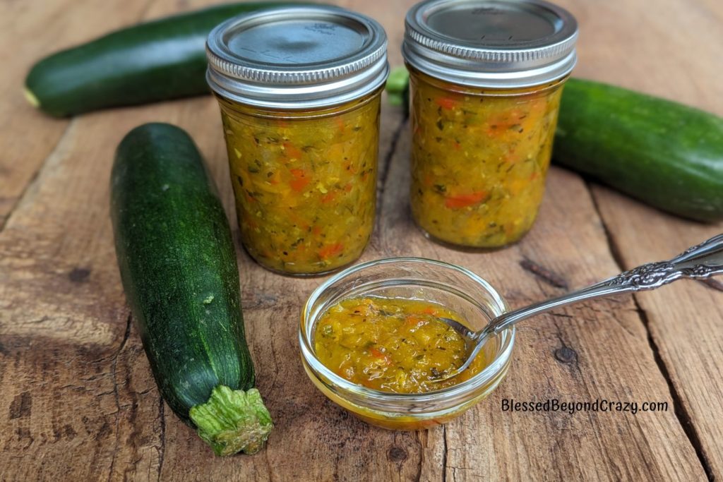 Two jars of zucchini relish pictured with fresh zucchini and tiny bowl of relish.