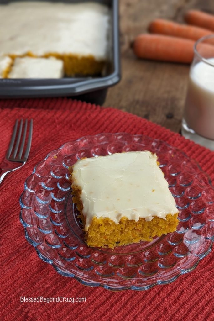 Slice of carrot bars with fresh carrots, a glass of milk, and pan of bars in the background.