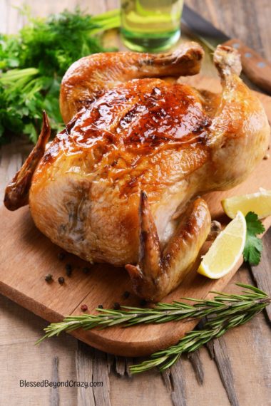 A whole roasted chicken resting on a wooden cutting board with slices of lemon and fresh rosemary sprigs.