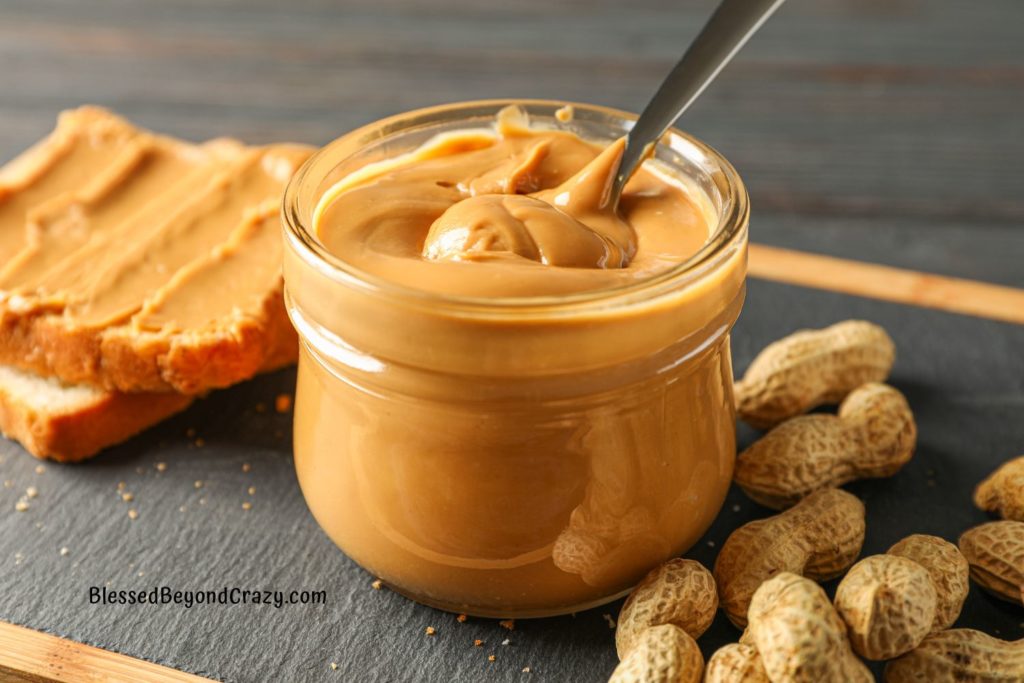 Photo of jar of creamy peanut butter with whole peanuts beside it and slices of bread with peanut butter.