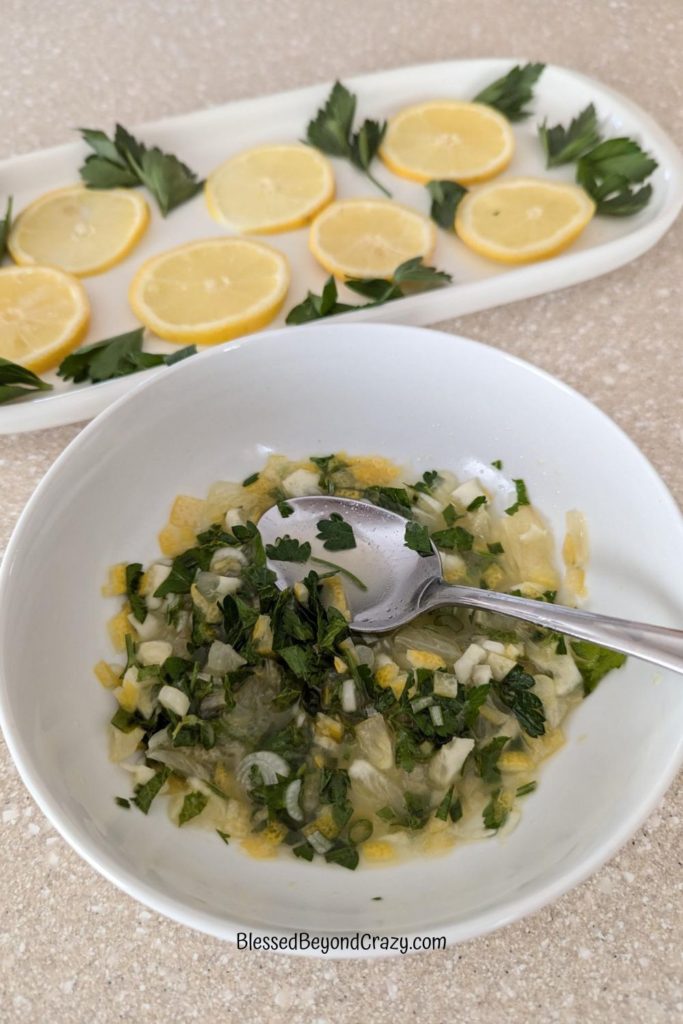 White bowl with mixed chopped herbs with platter in background with sliced lemons and fresh parsley.