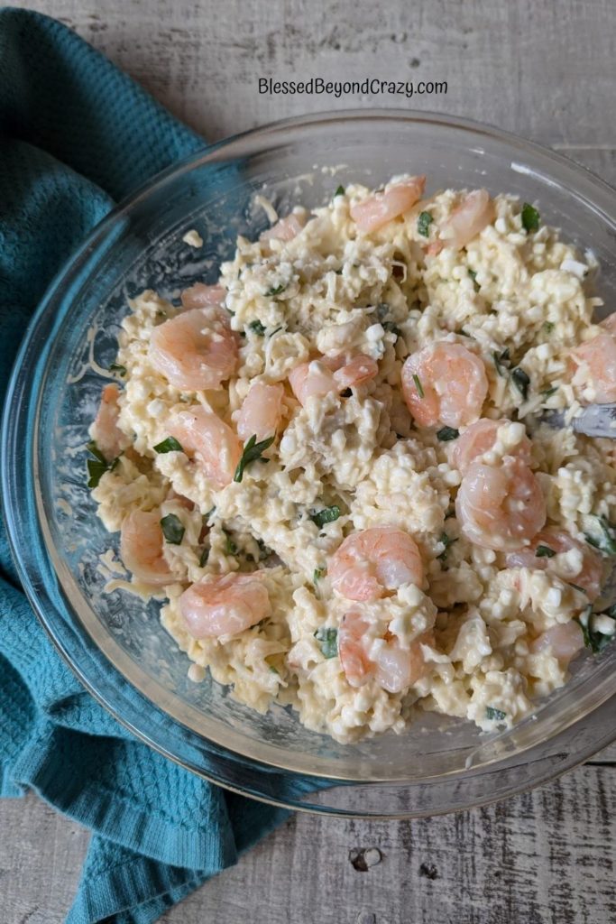 Overhead photo of glass bowl with seafood, cheese, and herbs.