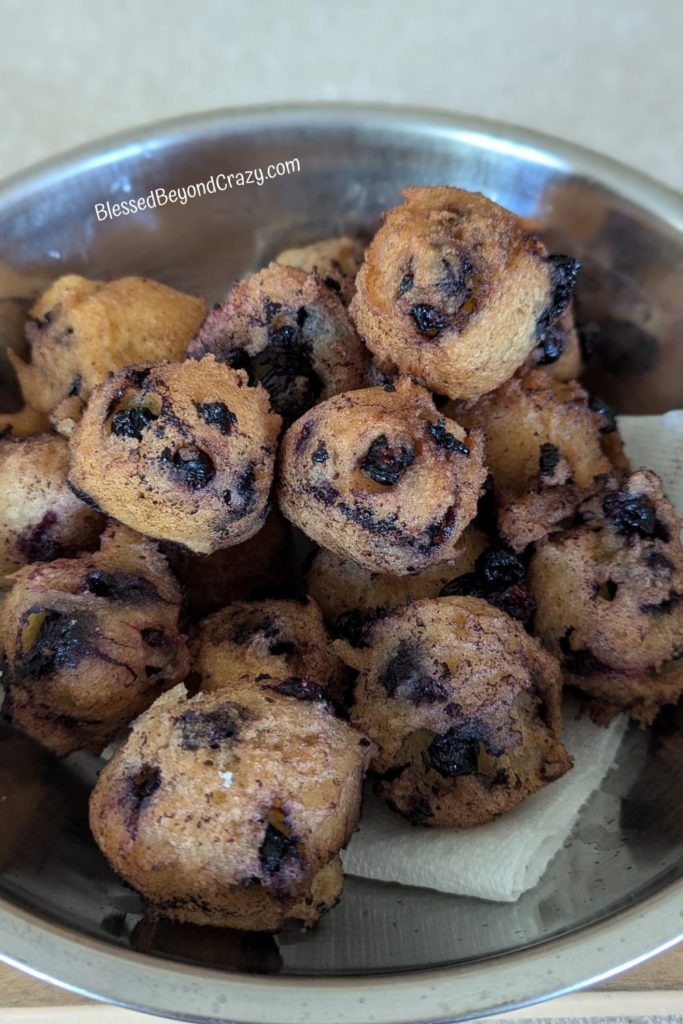 Freshly fried blueberry fritters cooling on paper towel inside a metal bowl.