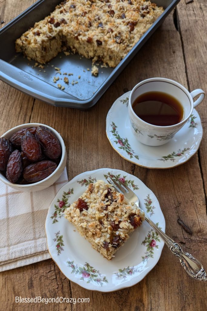 Overhead photo of individual serving of date coffee cake with a cup of hot tea, bowl of fresh dates, and remaining coffee cake in pan in background.