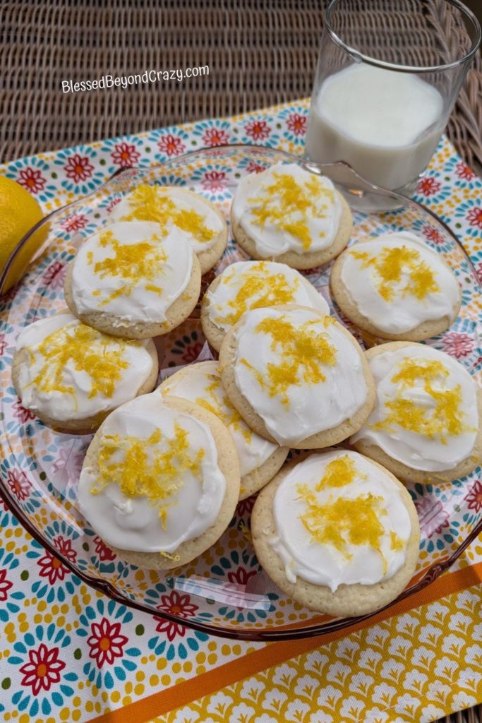 Plate of lemon drop cookies resting on colorful dish towel with glass of milk and fresh lemon.