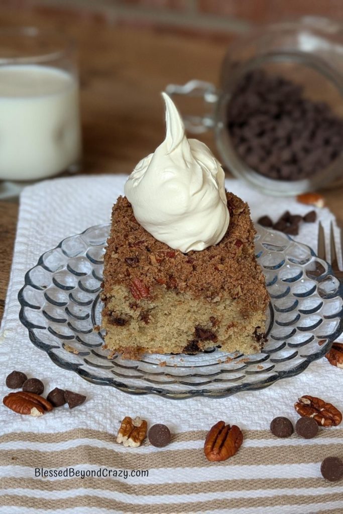 Individual serving of coffee cake with dollop of whipped topping, a glass of milk, and extra chocolate chips and pecans sprinkled on towel.