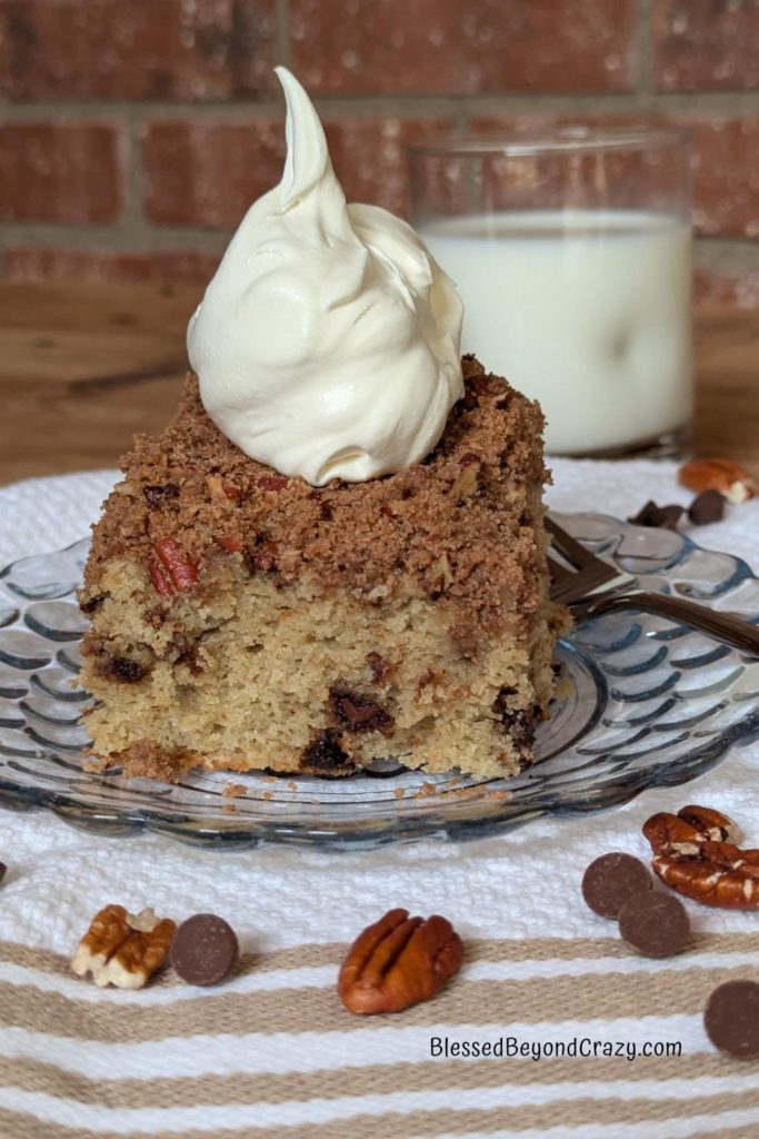 Close-up horizontal photo of serving of coffee cake garnished with whipped topping, a glass of milk, and chocolate chips and pecans.