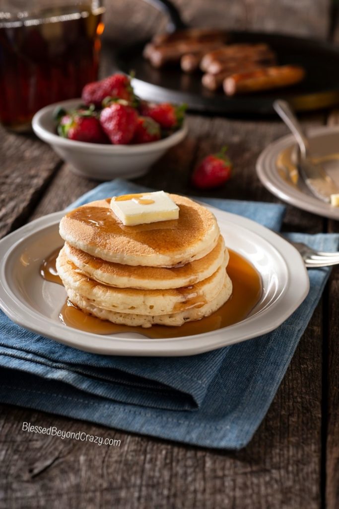 Vertical photo of stack of pancakes with bowl of fresh strawberries and sausage links in background.