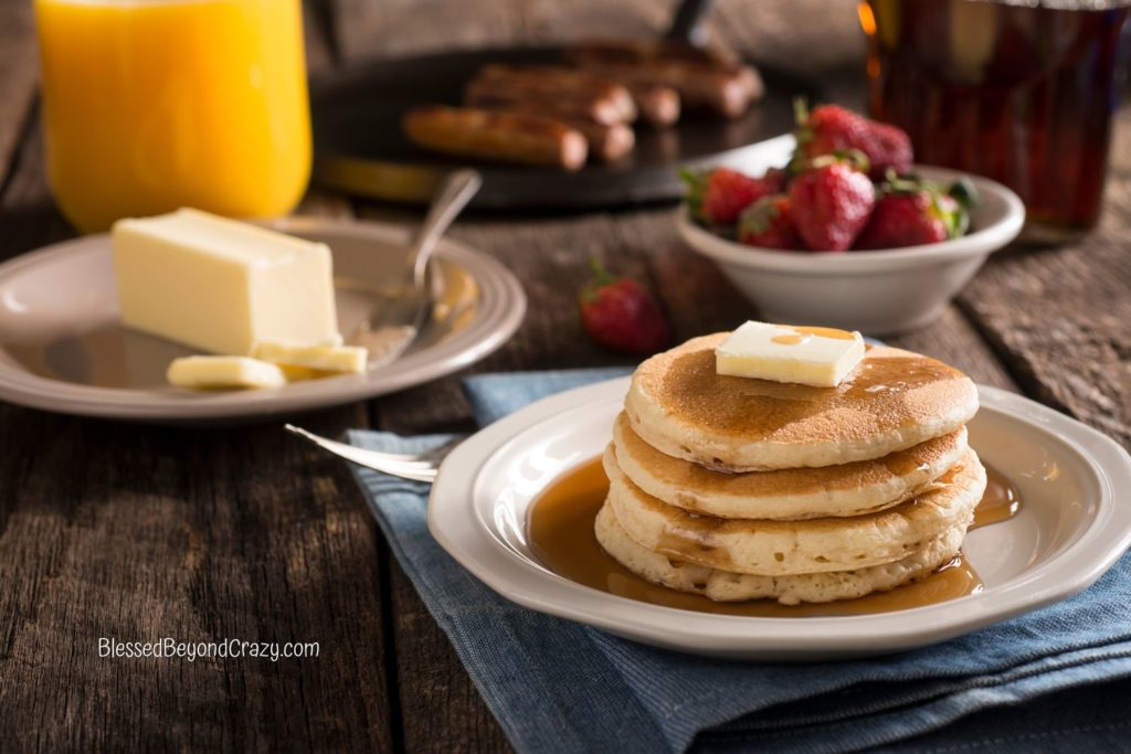 Photo with white plate with four pancakes, bowl of strawberries, griddle with sausage links, small plate with butter, and jar of maple syrup and pitcher of orange juice.