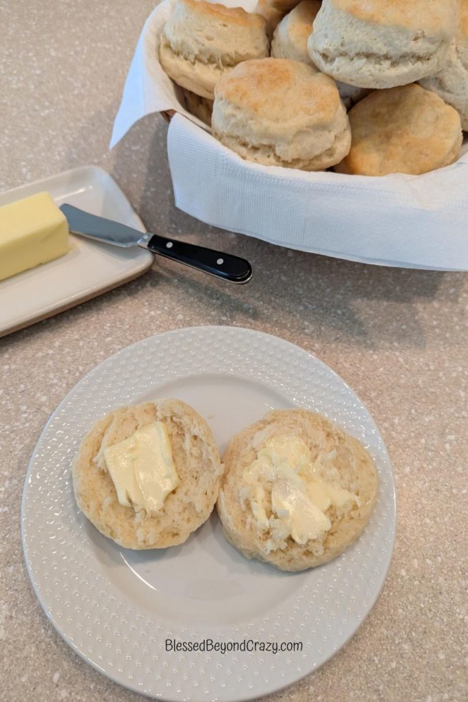 Overhead photo of a biscuit cut in half with butter on each section, along with a basket of biscuits and pad of butter.