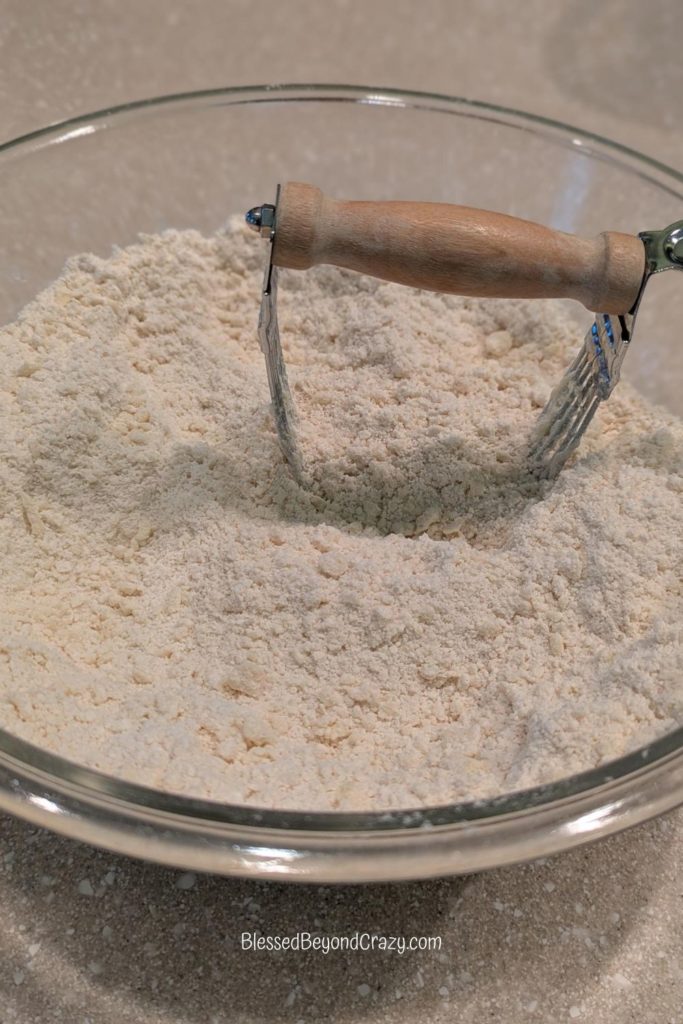 Clear glass mixing bowl with mixed flour and shortening with a pastry cutter.