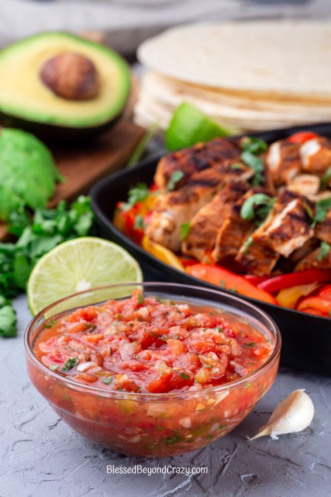 Vertical photo of bowl of salsa, fresh lime and avocado and skillet with fajita meat and vegetables